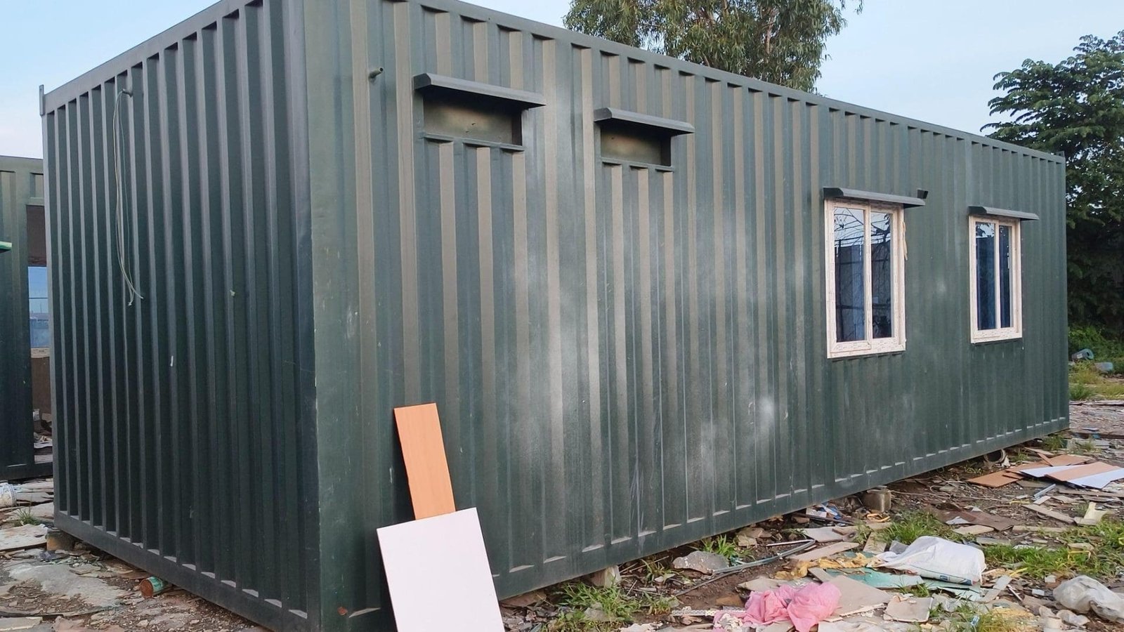 Large green portable cabin with multiple windows, placed on a construction site.